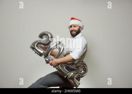 Ein Mann mit einem Bart in einem roten Samthut hält silberne Ballons in der Form von 2022 und hebt sein Bein. Party im neuen Jahr. Stockfoto