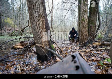 Eine Person im schwarzen Hoodie, die auf dem Sumpf im Herbstwald sitzt. Alte rostige Barrel an der Vorderseite. Assassin Cosplay Kostüm. Stockfoto