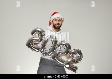 Ein unsicherer Mann in einem roten Samthut des Weihnachtsmanns hält silberne Ballons in Form von 2022. Ein Mann mit Bart auf einer Neujahrsparty. Stockfoto