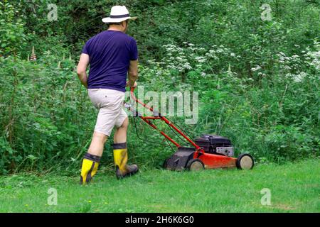 Rückseite des Mannes im Hut mäht grünes Gras mit Mähmaschine im Sommervorort Stockfoto