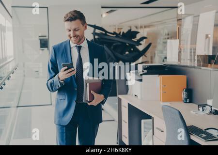 Junger, selbstbewusster Finanzier in formellem Anzug mit Laptop und Agenda, liest Nachrichten auf dem Smartphone Stockfoto