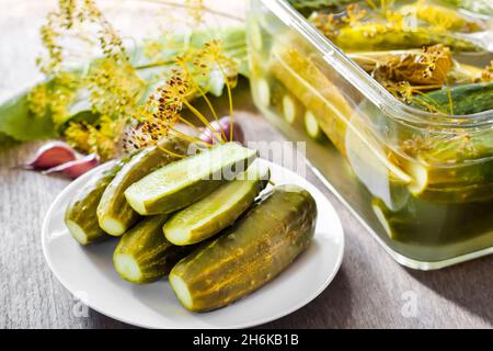 Eingelegte Gurken mit Kräutern und Gewürzen auf weißem Teller und Glasbehälter mit Gurken auf dem Hintergrund Stockfoto
