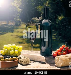 Flasche Rotwein, ohne Etikett und Glas voll Wein auf dem Tisch bei Sonnenschein mit Speisen, Käse, Oliven, Trauben und Brot Stockfoto