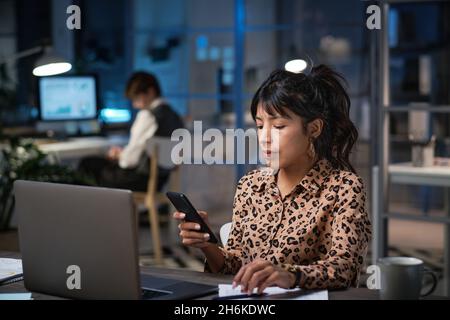 Geschäftsfrau, die vor dem Laptop am Tisch sitzt und während ihrer Arbeit im Büro eine Nachricht auf ihrem Mobiltelefon liest Stockfoto