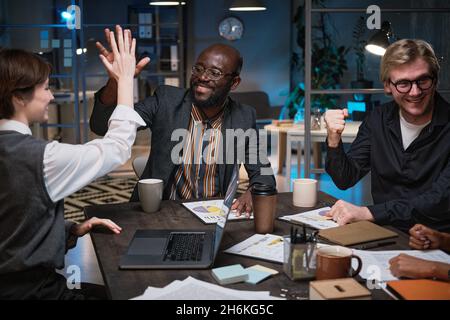 Glückliche junge Geschäftsleute, die während eines Geschäftstreffens im Büro bis spät in die Nacht am Tisch sitzen Stockfoto