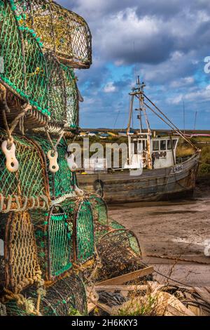 Stapel von Krabbenkäfigen im Vordergrund mit altem Fischerboot auf Schlamm bei Ebbe in Brancaster Staithe Harbour North Norfolk England Stockfoto