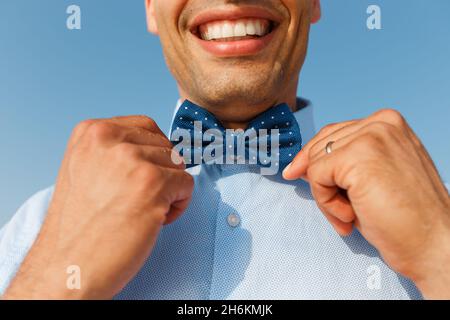Junger lächelnder Mann in blauem Hemd berührt die Fliege auf dem Hintergrund des blauen Himmels. Accessoires und Lifestyle für Herren Stockfoto