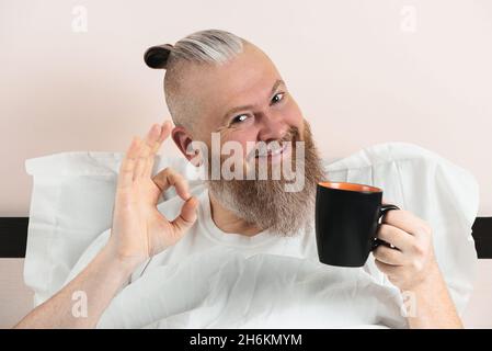 Ein glücklicher bärtiger Mann, der morgens Kaffee im Bett genoss und eine gute Geste zeigte. Lächelnder Hipster-Typ beginnt den Tag. Nahaufnahme. Stockfoto