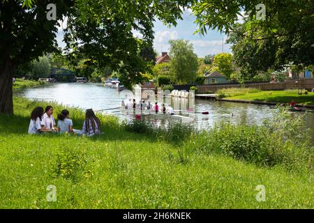 Mädchen, die zusammen unter einem Baum auf Gras neben dem River Cam in der Nähe von Cambridge sitzen, und Ruderer, die auf dem Downing College Eight am Fluss Cambridge in England rudern Stockfoto