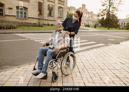 Junge Krankenschwester in schützender Gesichtsmaske, die sich während eines Spaziergangs in der Stadt um einen älteren behinderten Mann im Rollstuhl kümmert Stockfoto