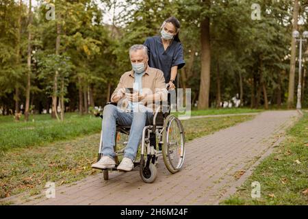 Junge Krankenschwester mit Gesichtsschutz und Schutzmaske, die sich während eines Spaziergangs im Park mit dem Smartphone um ältere behinderte Patienten im Rollstuhl kümmert Stockfoto