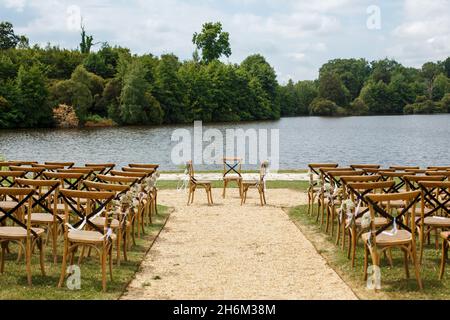 Hochzeitszeremonie auf dem Hintergrund des Sees. Reihen von Holzstühlen für Gäste, die im Garten des französischen Schlosses auf dem Gras stehen. Stockfoto