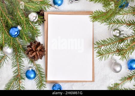 Eine leere Postkarte liegt auf einem Kraftumschlag im Schnee mit einem Weihnachtsbaum und blauen Glaskugeln. Banner, Einladung, Postkarte. Flach, Flach, Stockfoto