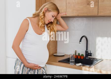 Schwanger blonde Frau fühlen sich schlecht in der Küche. Zukünftige Mutter krank, Kopfschmerzen und wollen Pille trinken Stockfoto