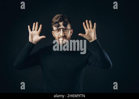 Junger bärtiger Hipster, der eine Sonnenbrille trägt, während er über einem isolierten schwarzen Hintergrund mit erhobenen Armen liegt Stockfoto