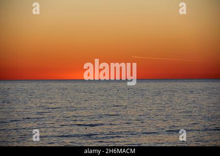 Die glühende Spur eines Flugzeugs in einem Abendhimmel nach Sonnenuntergang über einer ruhigen Meeresoberfläche Stockfoto