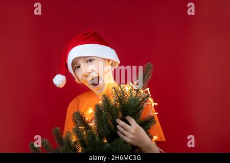 Der lustige Junge in der Weihnachtsmütze hält einen Weihnachtsbaum vor sich und lacht laut Stockfoto