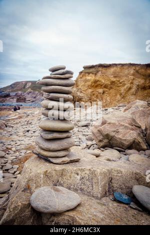 Am Dollar Cove Beach, Gunwalloe, Cornwall, England, liegen glatte Kieselsteine unter einem bewölkten Himmel mit zerklüfteten Klippen im Hintergrund. Stockfoto