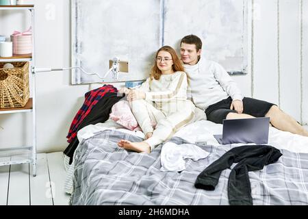 Halterung für Smartphone ist mit Klemme auf Rack in der Nähe des Bettes befestigt, junges verliebtes Paar schaut Video auf dem Telefonbildschirm, während es im Schlafzimmer auf dem Bett sitzt. Stockfoto