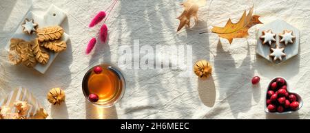 Panorama-Herbstbanner. Herbstdekorationen - Preiselbeere, Kekse. Tee und trockene Eichenblätter. Flach liegend, Draufsicht auf unfarbigem Textil. Herbsthintergrund Stockfoto