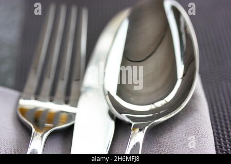 Metallsuppenlöffel, Messer, Gabel liegen auf einer grauen Serviette auf einer schwarz-grauen Oberfläche Draufsicht. Tischeinrichtung, Tuch im Restaurant, Café, in der heimischen Küche. Grau Stockfoto