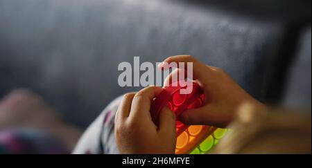 Kleines Kind spielt mit Regenbogen Pop-it Fidget Gadget. Beliebtes Anti-Stress-Spielzeug aus Silikon. Jugendkultur und Konzept der psychischen Gesundheit. Modisches, flexibles Spielzeug Stockfoto