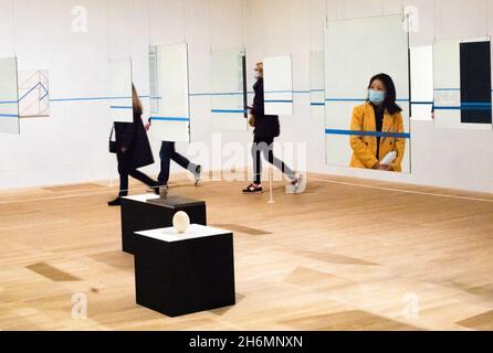 Eine asiatische Frau mit einer OP-Maske wird in einem Spiegel gesehen, während andere in einer Galerie vorbeigehen, die eine Kunstinstallation in der Tate Modern zeigt Stockfoto