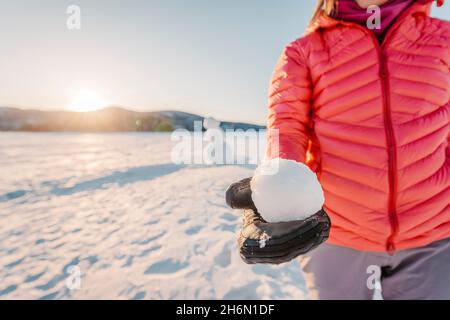 Winter Outdoor Active Lifestyle Konzept. Mädchen hält Schneeball an verschneiten Tag mit Schneemann im Hintergrund auf gefrorenen See bei schönen Winter Sonnenuntergang Stockfoto