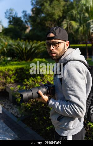 Ein junger bärtiger Mann mit einer Mütze, der Fotos in der Natur mit Bäumen und Sträuchern im Hintergrund fotografiert Stockfoto