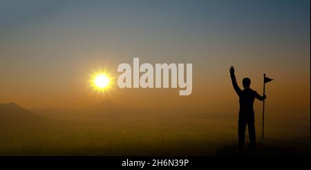 Geschäftsmann hält Flagge und hebt Hand auf den Berg mit Sonnenuntergang. Silhouette des männlichen erfolgreichen Unternehmers Blick auf den Sonnenuntergang. Stockfoto