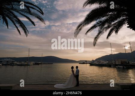 Tivat, Montenegro - 17.06.19: Braut und Bräutigam halten sich die Hände, während sie bei Sonnenuntergang auf dem Pier vor der Kulisse der Berge und der Bucht stehen Stockfoto