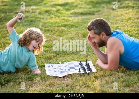 Vater und Sohn spielen Schach und liegen auf dem Rasen im Rasenpark. Vatertag, Liebesfamilie, Elternschaft, Kindheitskonzept. Stockfoto