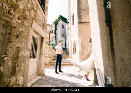 Sibenik, Kroatien - 05.06.17: Bräutigam, die Arme auf der Brust gefaltet, steht auf der Straße in der Nähe der alten Häuser. Braut geht um die Ecke des Hauses Stockfoto