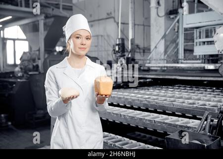 Porträt einer Bäckerin, die Teig und frisches Brot in den Händen auf dem automatischen Hintergrundförderer der Bäckerei hält. Stockfoto