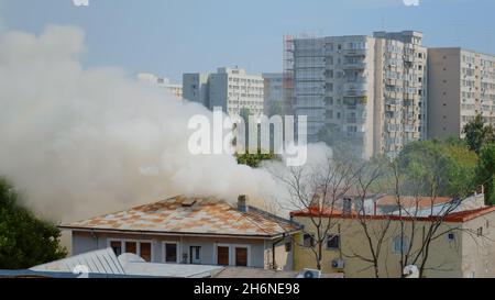 Ansicht der Feuerwehrleute, die das Feuer aus dem brennenden Haus mit Wasser löschen. Rauch und Flammen, die durch ein Feuer vom Dach des Gebäudes entstehen und gefährlichen Smog und Rauch in der Nachbarschaft der Stadt ausbreiten. Stockfoto