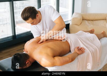 High-Angle-Ansicht des professionellen männlichen Masseurs mit starken Händen massieren Schulterblätter und Rücken des muskulösen Sportmanns auf dem Bauch liegen. Stockfoto