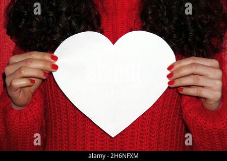 Lockiges Haar Mädchen in roten Pullover hält weißes Herz mit Platz für Text. Valentinstag-Konzept Stockfoto