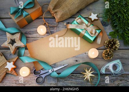 Geschenkverpackung mit braunem Papier und türkisfarbenem Band, Scheren, Dekorationssternen und brennenden Kerzen auf einem grauen rustikalen Holztisch, Kopierraum Stockfoto