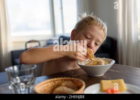 Kleiner Junge, der Spaghetti mit Händen im Wohnzimmer isst Stockfoto