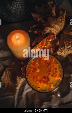 Brennende Kerze und eine große Tasse heißen orangefarbenen Sanddorntee in einem gemütlichen herbstlichen Stillleben mit trockenen Blättern. Stockfoto