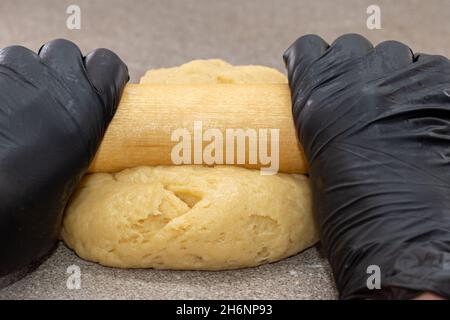 Die Hände des Küchenchefs in schwarzen Handschuhen Rollen den Teig mit einem Nudelholz auf einem grauen Tisch aus der Nähe aus. Kochteig für Kuchen, Pizza, Kekse. Hausgemachtes bakin Stockfoto