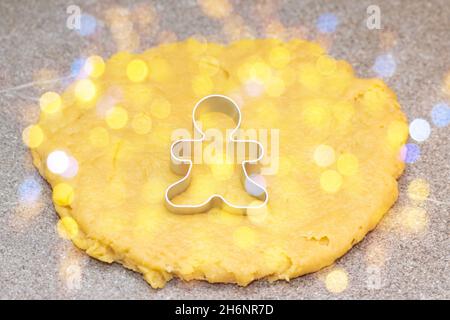 Form zum Schneiden von Lebkuchenmännern auf rohem, gewalztem Teig auf grauem Hintergrund mit goldenem, rundem Bokeh. Der Prozess der Herstellung von magischen Weihnachtsplätzchen bei Stockfoto