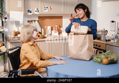 Lächelnde Frau, die das Essen aus der Papiertüte am Tisch in der Küche herausnahm und Essen für eine ältere Frau lieferte Stockfoto