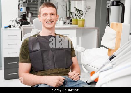 Junger Mann mit Zahnspangen an den Zähnen besucht Zahnarztpraxis. Lächelnder Mann mit schwarzem Lätzchen, sitzt auf dem Zahnarztstuhl, in einem gut ausgestatteten Büro. Konzept der zeitgenössischen Zahnheilkunde Stockfoto