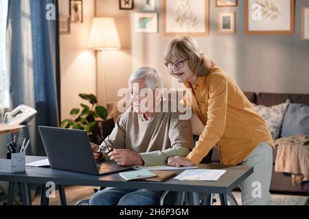 Eine ältere Frau in einer Brille half ihrem Mann, den Laptop zu benutzen, während sie zu Hause am Tisch saß Stockfoto