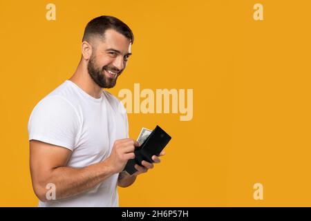 Schlauer, lächelnder Europäer mittleren Alters in weißem T-Shirt, der Geld in die Brieftasche legte und die Kamera betrachte Stockfoto