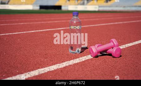 Langhanteln und Flasche auf der Laufstrecke. Sportgerät und -Ausrüstung. Kurzhanteln auf der Rennstrecke im Stadion. Hydratation nach Fitness. Gesunder, ausgewogener Lebensstil Stockfoto