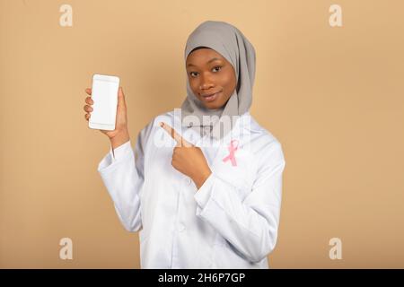 Schöne muslimische Religion Ärztin mit rosa Bogen gegen Krebs zeigt einen leeren Smartphone-Bildschirm isoliert auf einem gelben Hintergrund Stockfoto