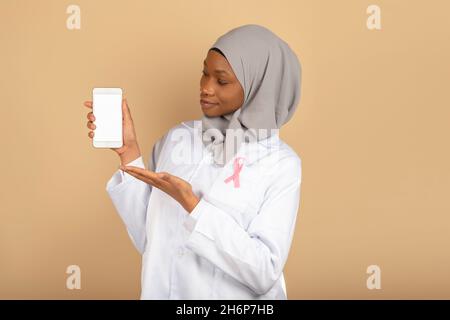 Schöne muslimische Religion Ärztin mit rosa Bogen gegen Krebs zeigt einen leeren Smartphone-Bildschirm isoliert auf einem gelben Hintergrund Stockfoto
