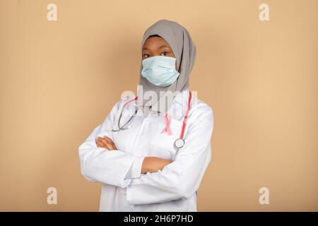 Porträt einer afroamerikanischen Ärztin, die mit gekreuzten Armen eine OP-Maske, einen Schal und einen weißen Mantel trägt. Stockfoto